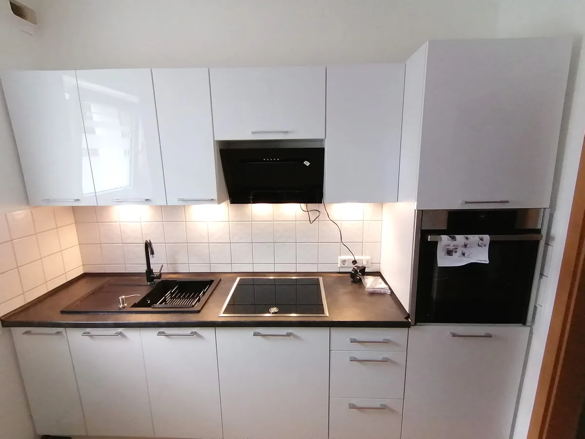 Weiße Küchenzeile mit schwarzer Spüle, Kochfeld im Edelstahlrahmen und Backofen im Hochschrank