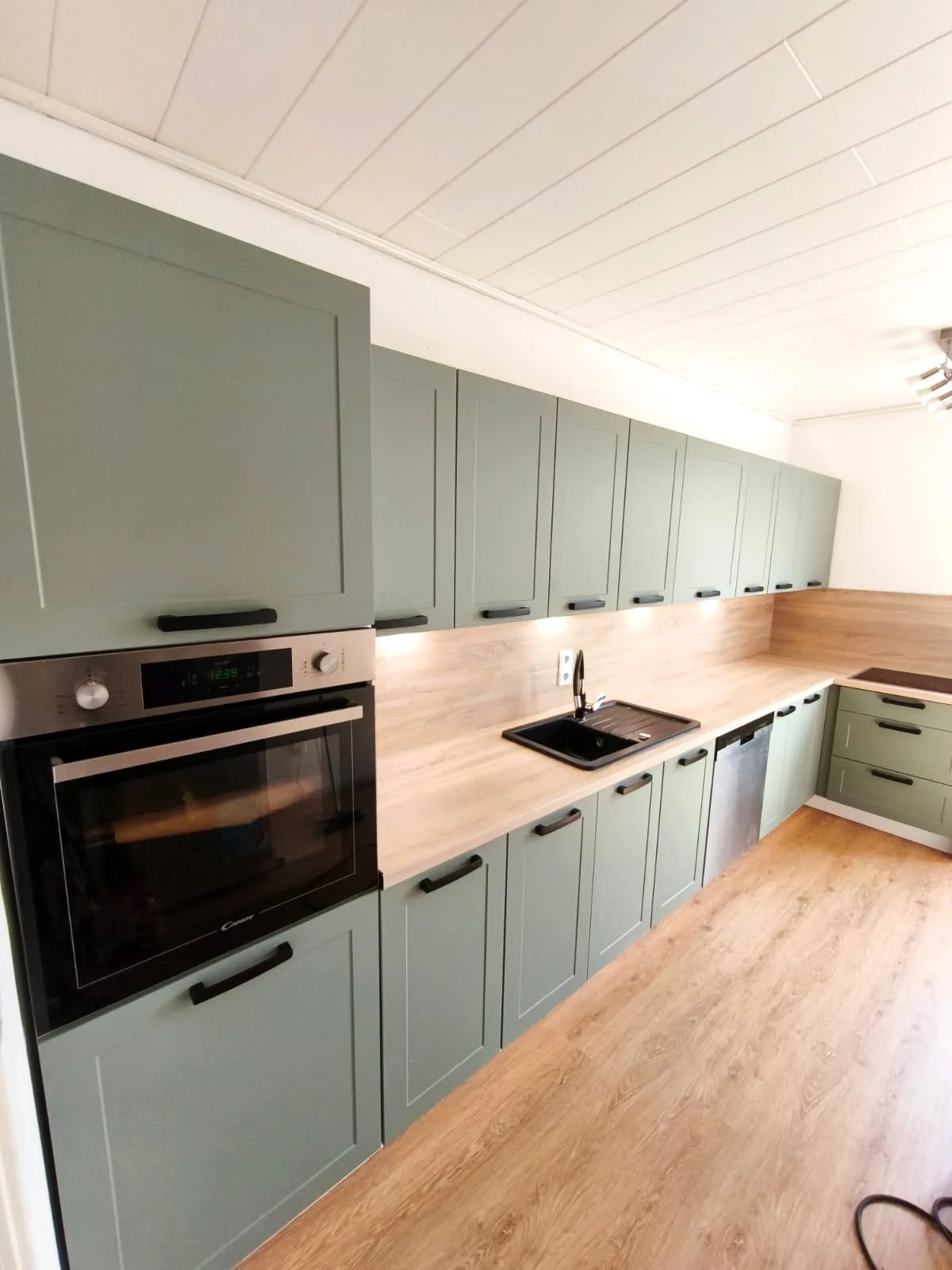 Lange Küche mit salbeigrünen Rahmenfronten, heller Holzdekor-Arbeitsplatte und Backofen im Hochschrank